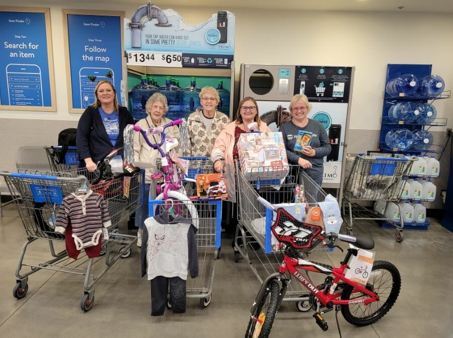 Five women stand behind three shopping carts. Inside the shopping carts are kids' clothing, toys and bicycles that they are purchasing.