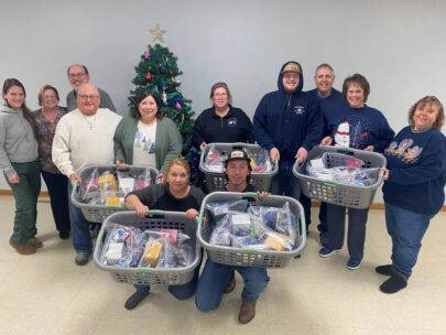 12 people pose for the picture with five laundry hampers full of gift bags. There is a Christmas tree behind the group.
