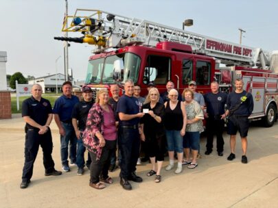 15 people are standing together, posing for the photo. They are outdoors, standing in front of a fire truck that says "Effingham Fire Dept." Standing in the front and middle of the group are four women, who are handing over a gift card to one of the firefighters.