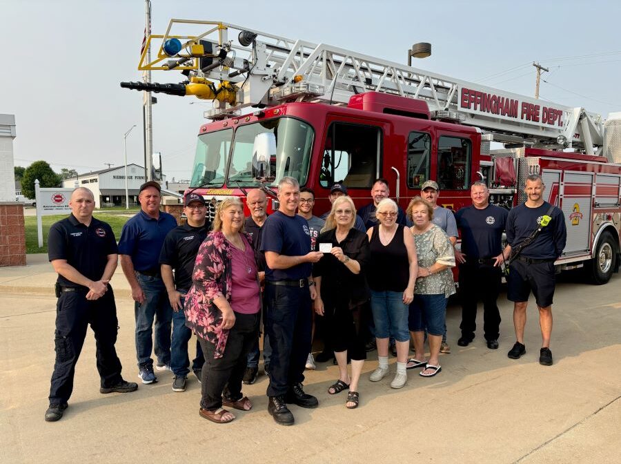 15 people are standing together, posing for the photo. They are outdoors, standing in front of a fire truck that says "Effingham Fire Dept." Standing in the front and middle of the group are four women, who are handing over a gift card to one of the firefighters.
