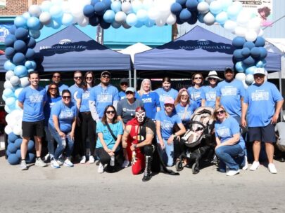 A group of about 20 WoodmenLife associates stand together for the photo. They are outside in the sunshine, and behind them are blue WoodmenLife-branded tents and a blue-and-white balloon arch. Everyone is wearing a blue WoodmenLife t-shirt. A person in the middle of the group is wearing a black-and-red luchador costume.