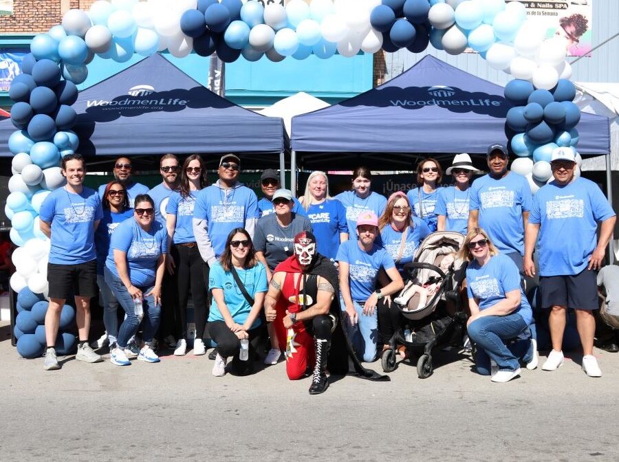 A group of about 20 WoodmenLife associates stand together for the photo. They are outside in the sunshine, and behind them are blue WoodmenLife-branded tents and a blue-and-white balloon arch. Everyone is wearing a blue WoodmenLife t-shirt. A person in the middle of the group is wearing a black-and-red luchador costume.