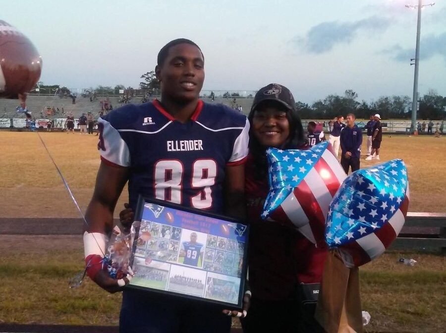 Photo shows a woman and a young man. The young man is wearing a football uniform and holding a framed picture; the woman is wearing a baseball hat and holding red-white-and-blue balloons. Behind them is a football field.