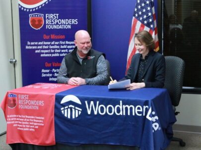 Two people — a man and a woman — sit at a table. A red table cloth in front of the man has the First Responders Foundation logo, and a blue table cloth in front of the woman has the WoodmenLife logo. The woman is signing a piece of paper.