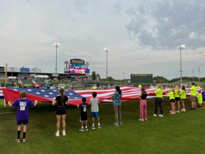 A group of about 40 people are surrounding and holding a 30-foot-by-60-foot American flag. They are on the outdoor field of the Union Omaha soccer team.