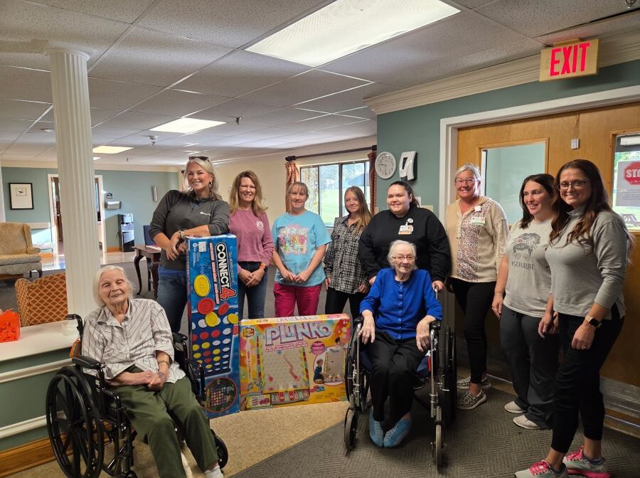 10 people pose for the photo, two of whom are elderly women in wheelchairs. In the middle of the group are a large box containing a Connext 4 game and a large box containing a Plinko game.