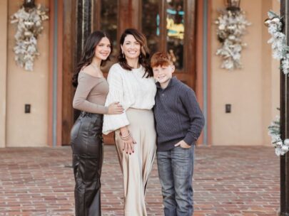 Photo shows a woman in her 30s, flanked by a daughter and a son. The daughter is a teenager, and the son is a pre-teen. The three people are standing outside on a brick walkway, and a building's doors are behind them.