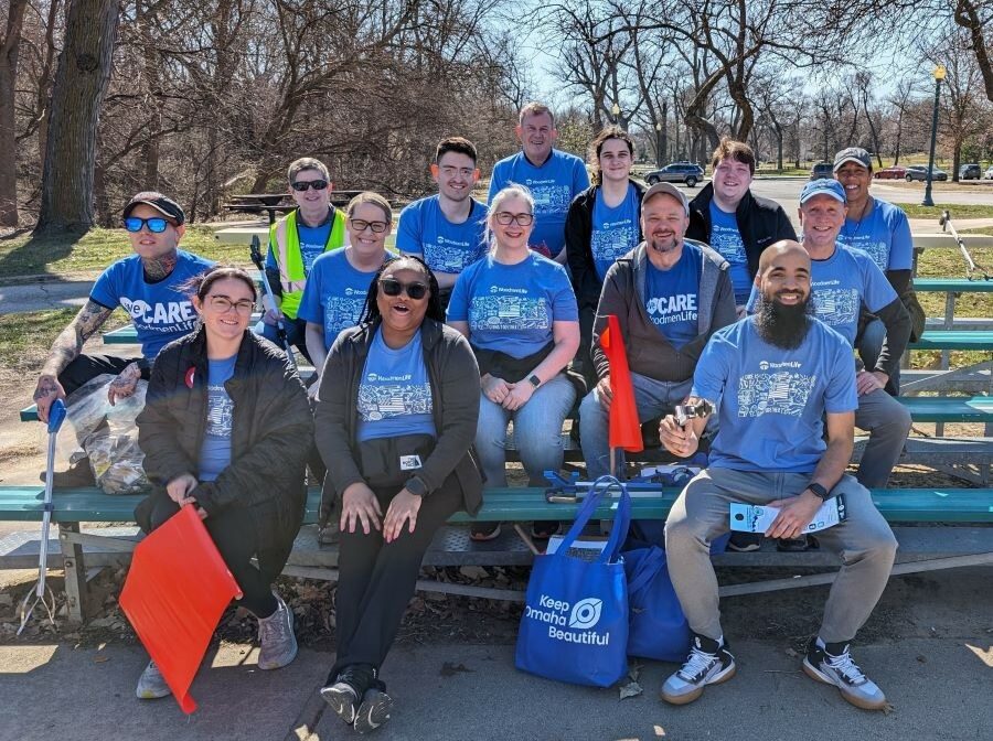 A group of 14 WoodmenLife associates — volunteers for Keep Omaha Beautiful — are seated on outdoor bleacher seats. They are all wearing blue WoodmenLife-branded t-shirts. Some people are holding bags and "grabbers" used to pick up litter.