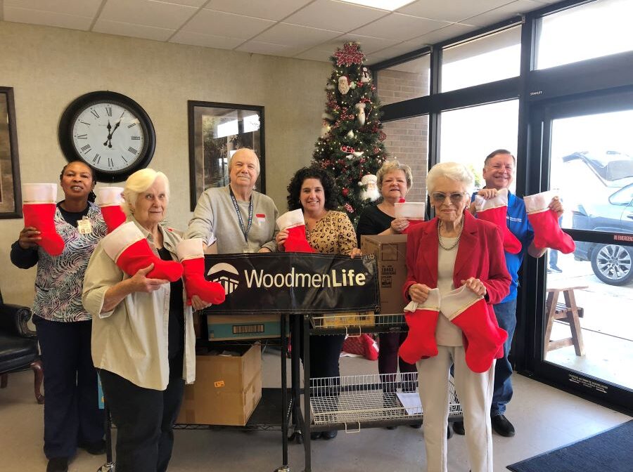 A group of seven people pose for the picture. They are all holding up Christmas stockings, which are red with a white band at the top. Behind them is a Christmas tree.