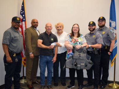 Seven people stand together for the photo, three of whom are wearing police uniforms. Two of the people in the middle are holding a check, and two other people are holding up a bulletproof vest. Behind them are an American flag and a state flag on flagpoles.