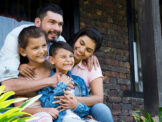 Stock photo shows a family, which includes a man, a woman, a young girl, and a young boy. They are sitting together on the front porch of a home, sitting close together, smiling, and embracing.