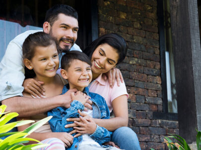 Stock photo shows a family, which includes a man, a woman, a young girl, and a young boy. They are sitting together on the front porch of a home, sitting close together, smiling, and embracing.