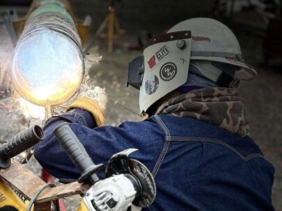 Photo shows a person welding. The photo is shot from over their shoulder as they work. They are wearing a protective mask, long-sleeve shirt, and gloves.