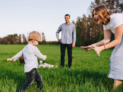 Stock photo shows a man, woman and young boy. They are outside in a grassy field. The woman and boy are playing in the foreground, and the man is looking on in the background. There are trees farther behind them.