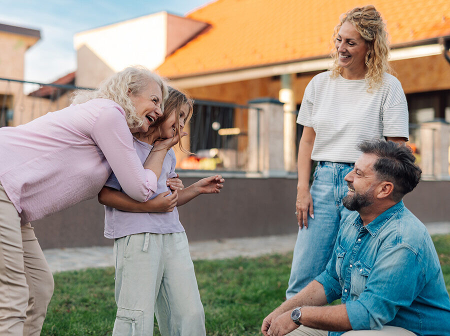 Stock photo shows four people. An older woman smiling and embracing a young girl on the left, and an adult man and woman smiling and looking on from the right. They are standing outside.
