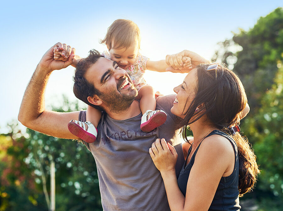 Image is a stock photo of a man, woman, and young child. The child is sitting on the man's shoulders, and both the man and woman are smiling up at the child. They are outdoors, and the sun and green trees are behind them.