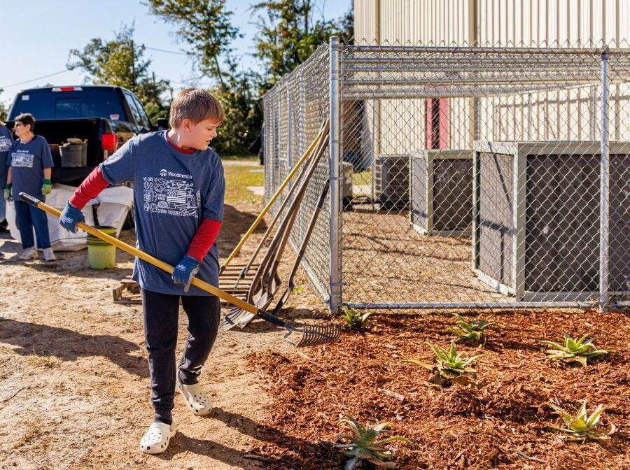 A boy uses a garden hoe to spread around newly laid mulch. He is wearing a blue WoodmenLife-branded Giving Together t-shirt.