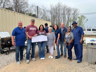 Eight people stand together for the photo. Two people in the middle, both wearing mauve-colored shirts that say "HOPE," are holding an oversized WoodmenLife-branded check made out to "HOPE Campaign" in the amount of $500. The group is standing outside on a gravel walkway with trees behind them.