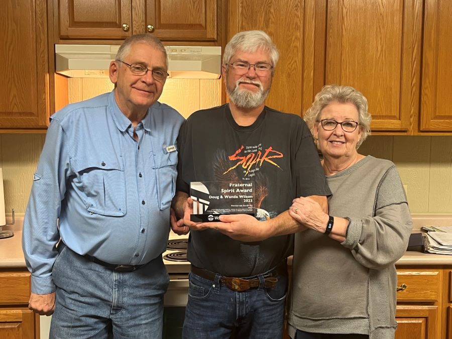 Two men and a woman stand together posing for a picture. The man in the middle is holding a glass award that says "Fraternal Spirit Award, Doug & Wanda Wilson, 2023."