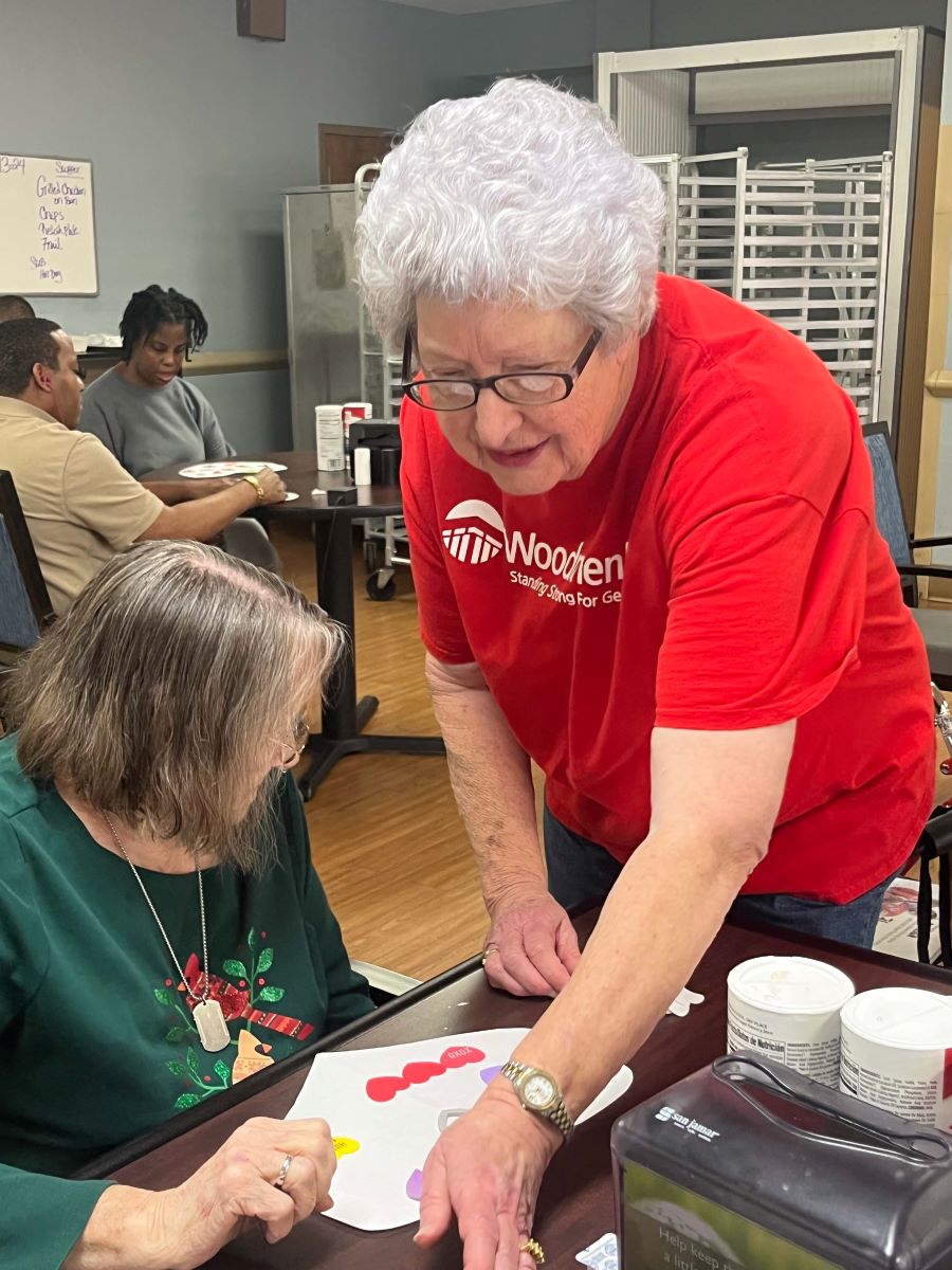 A woman in a red WoodmenLife T-shirt is standing and helping another woman, who is seated, with a craft project.