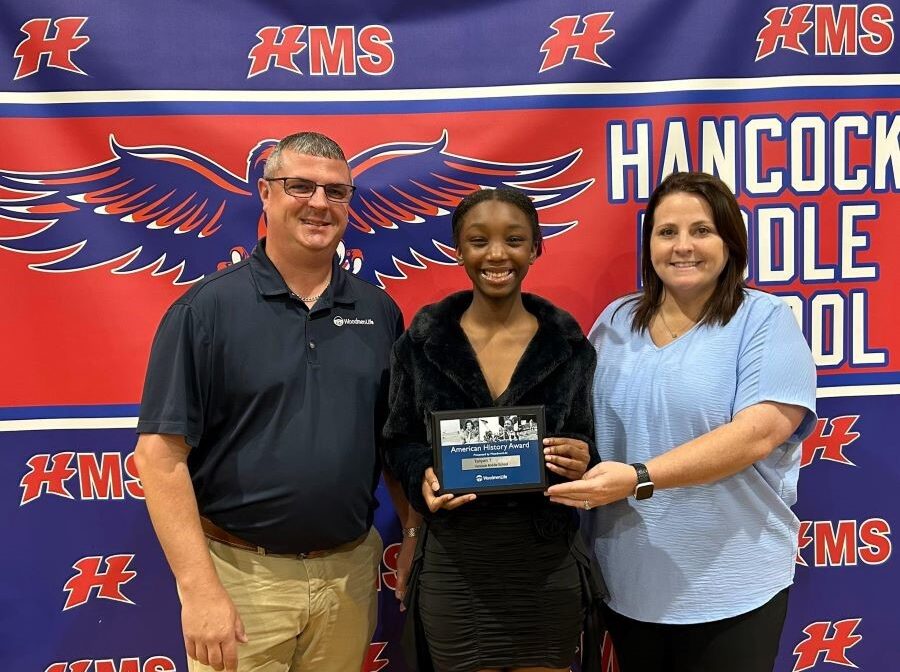 A man and a woman flank a middle school student, who is holding a small plaque. Behind them is a school-branded backdrop that is primarily blue and red, and says "Hancock Middle School."