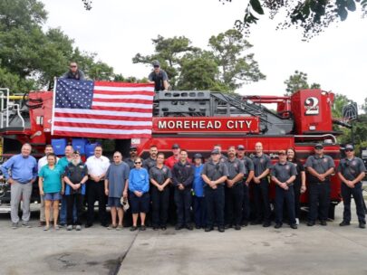 A total of 25 people are shown in the photo. 23 of the people are standing in a row in front of a fire truck, and two people are atop the fire truck, holding open a U.S. flag.