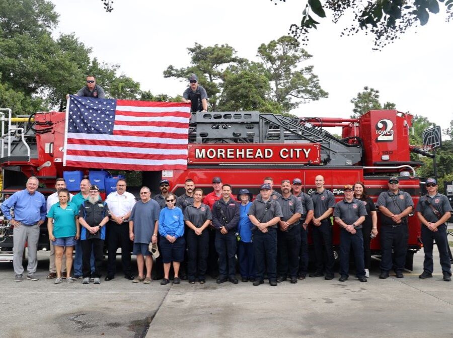 A total of 25 people are shown in the photo. 23 of the people are standing in a row in front of a fire truck, and two people are atop the fire truck, holding open a U.S. flag.