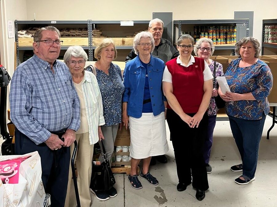 A group of eight people stand together. Behind them are shelves with nonperishable food.