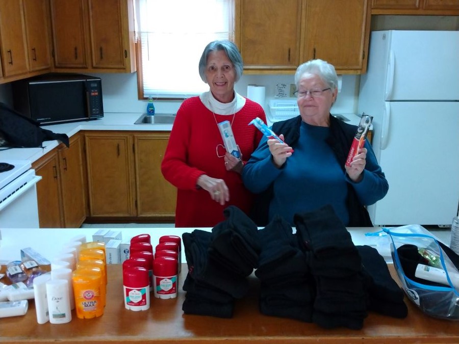 Two women stand behind a counter filled with donations — including deodorant and toothbrushes — for a local homeless shelter.