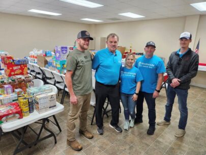 Five people stand together for a photo. They are all wearing WoodmenLife-branded shirts or hats. Behind them are multiple long tables stacked with supplies, like diapers and shelf-stable food.