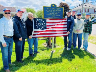 Seven people stand together holding a flag. The commemorative flag has 13 stars in a circle around the number 250 in upper-left blue square. The group is standing in front of a sign that says "East Battery. On this hill was encamped the artillery of the American Army under Gen. George Izard. 1814. 2026 by WoodmenLife Chapter 462." There is green grass below them, and houses and trees behind them.