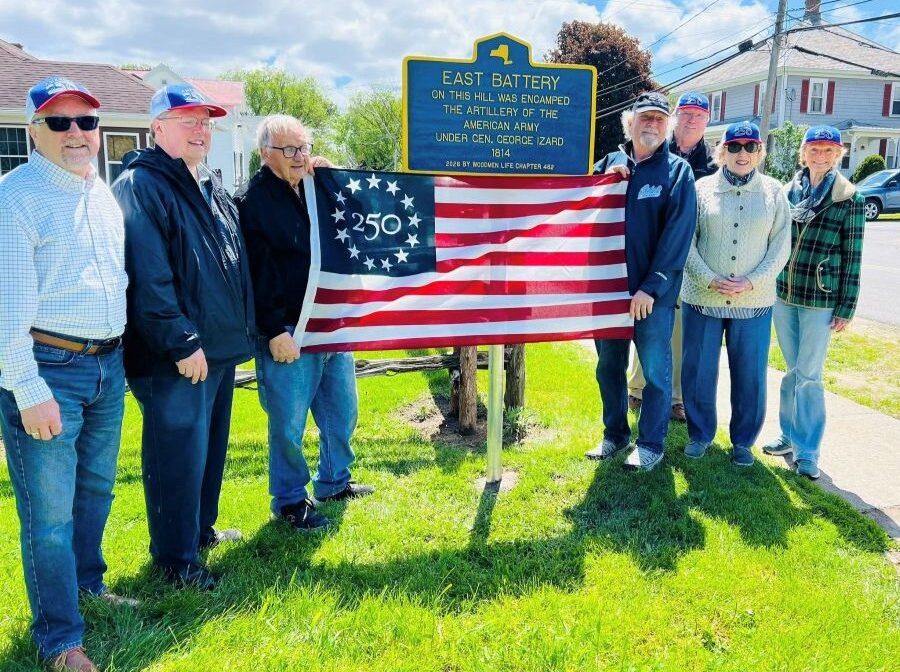 Seven people stand together holding a flag. The commemorative flag has 13 stars in a circle around the number 250 in upper-left blue square. The group is standing in front of a sign that says "East Battery. On this hill was encamped the artillery of the American Army under Gen. George Izard. 1814. 2026 by WoodmenLife Chapter 462." There is green grass below them, and houses and trees behind them.