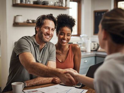 Image is a stock photo of a man and woman. They are sitting at a table, and the man is reaching out to shake hands with the person across from them. There are sheets of paper and a laptop on the table in front of the couple.