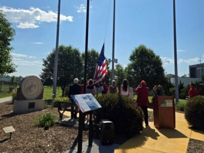 A group of local dignitaries stand facing a flag as it's raised on a flag pole. The group is standing outside on a sunny day.