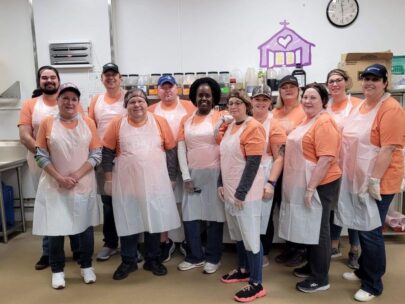 12 WoodmenLife associates pose for a photo while volunteering at Open Door Mission. All are wearing orange T-shorts, plastic aprons and either baseball hats or hairnets. On the wall behind the group is the Open Door Mission logo.