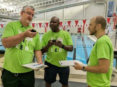 Three volunteers with Special Olympics are standing beside a swimming pool, looking closely at stopwatches. They are all wearing lime green shirts indicating they're volunteers.