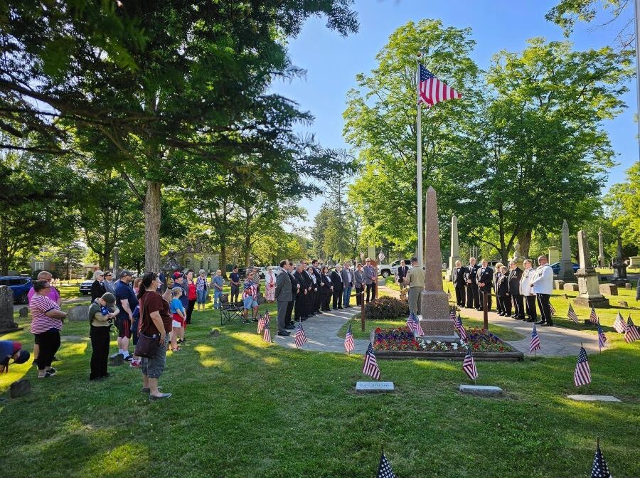 A group of people are gathered around a flag pole in the middle of a cemetery. The flag is at the gravesite of of Francis Bellamy, the author of the Pledge of Allegiance.