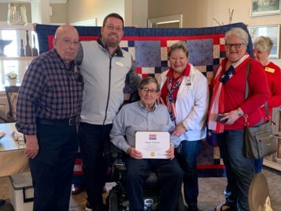 Five people stand together for the photo. The man in the middle, who is seated in a wheelchair, is holding a certificate. A quilt is being held up behind the group.