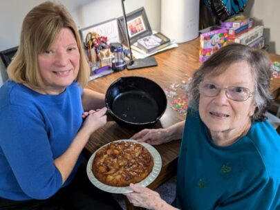 Executive Vice President & Chief Risk Officer Shawn Bengtson and her mother sit at a table. Shawn is holding an empty cast iron skillet, and her mother is holding the peach upside-down cake on a serving plate.