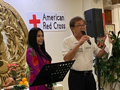 A man and a woman are standing. The man, Recruiting Sales Manager Chris Nguyen, is holding a microphone as he announces that WoodmenLife is donating $10,000 to the American Red Cross to help with its Maui wildfires recovery effort. A sign on the wall behind them says "American Red Cross."