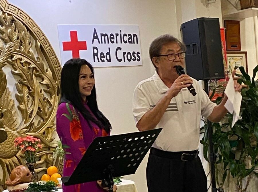 A man and a woman are standing. The man, Recruiting Sales Manager Chris Nguyen, is holding a microphone as he announces that WoodmenLife is donating $10,000 to the American Red Cross to help with its Maui wildfires recovery effort. A sign on the wall behind them says "American Red Cross."