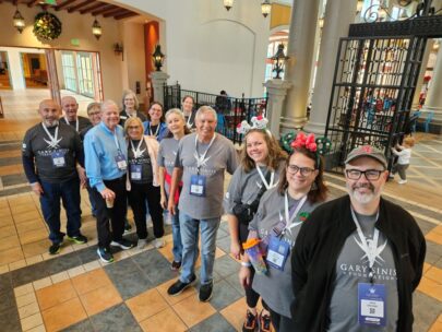 A group of 13 people stand together for the photo. They are wearing dark gray T-shirts that have the Gary Sinise Foundation logo on the front.