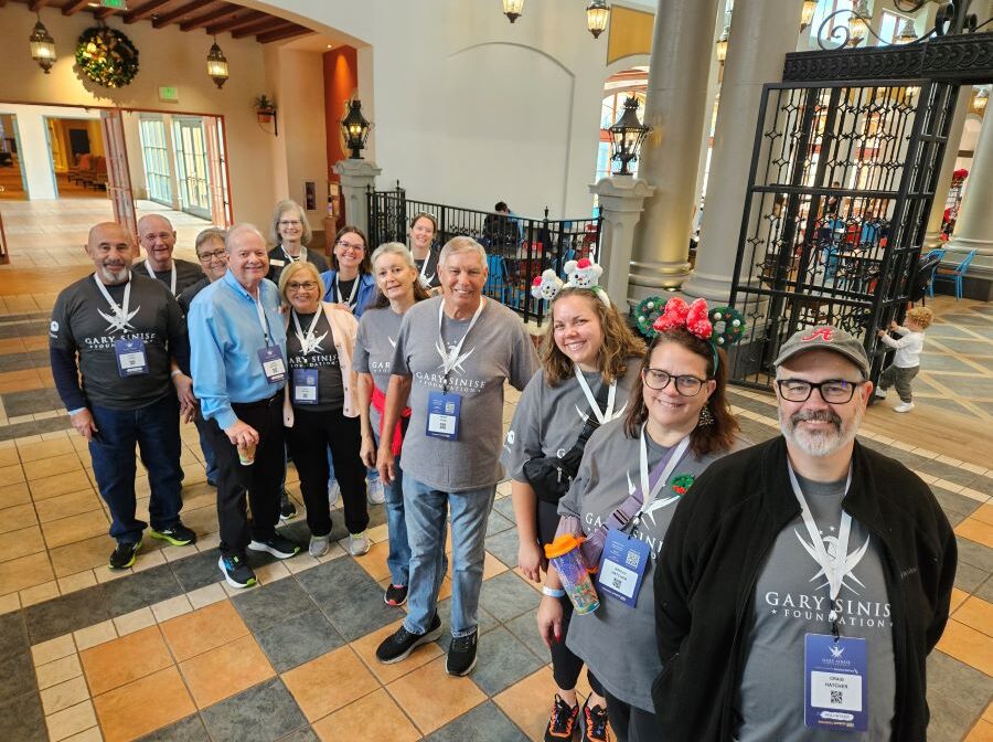 A group of 13 people stand together for the photo. They are wearing dark gray T-shirts that have the Gary Sinise Foundation logo on the front.