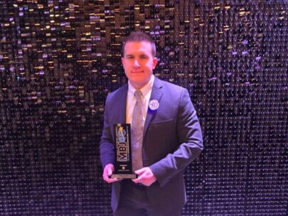 Photo shows a man in a suit holding an award. Behind him is a shiny, textured backdrop.