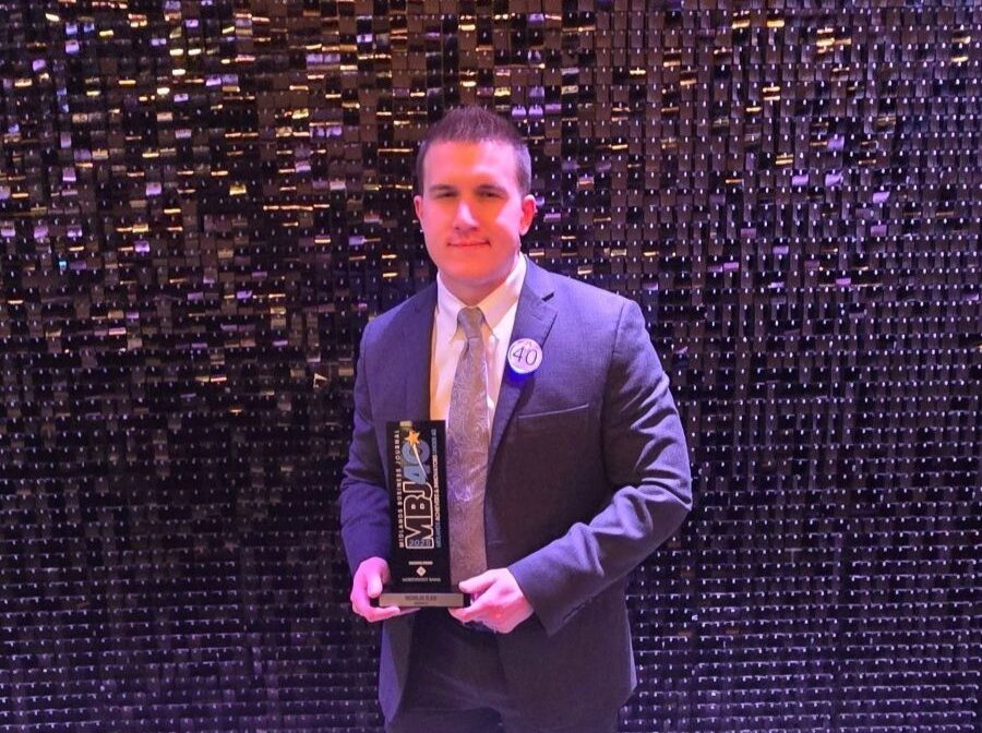Photo shows a man in a suit holding an award. Behind him is a shiny, textured backdrop.