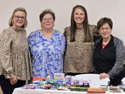 Four women stand together in the photo. In front of them is a table with school supplies, including glue, Sharpies, and markers.