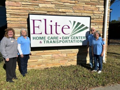 Four people flank a sign on the outside of a building. The sign says "Elite Home Care, Day Centers & Transportation."