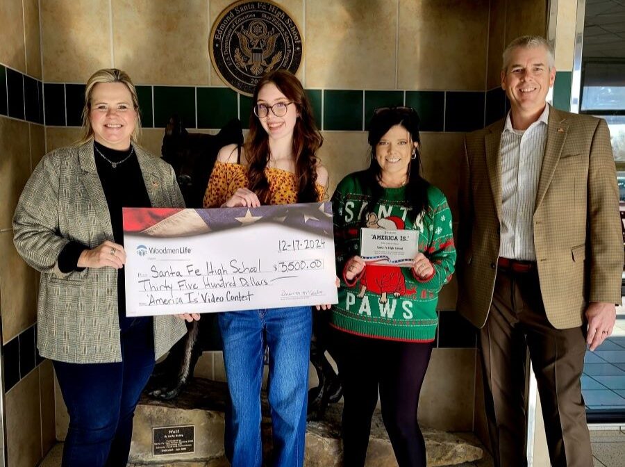 Four people pose with an oversized WoodmenLife-branded check. The check is made out to Santa Fe High School in the amount of $3,500. The woman second from the right is holding a small plaque that says "America Is ..." at the top.