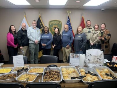 A group of 10 people stand in a row. In front of them is a table filled with aluminum pans of breakfast foods: cinnamon rolls, breakfast sandwiches, eggs, sausage, and potatoes. Behind them are flags and a Sheriff's Department seal on the wall.