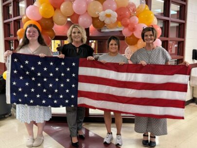 Four women stand holding up an American flag. Behind them is a balloon arch.